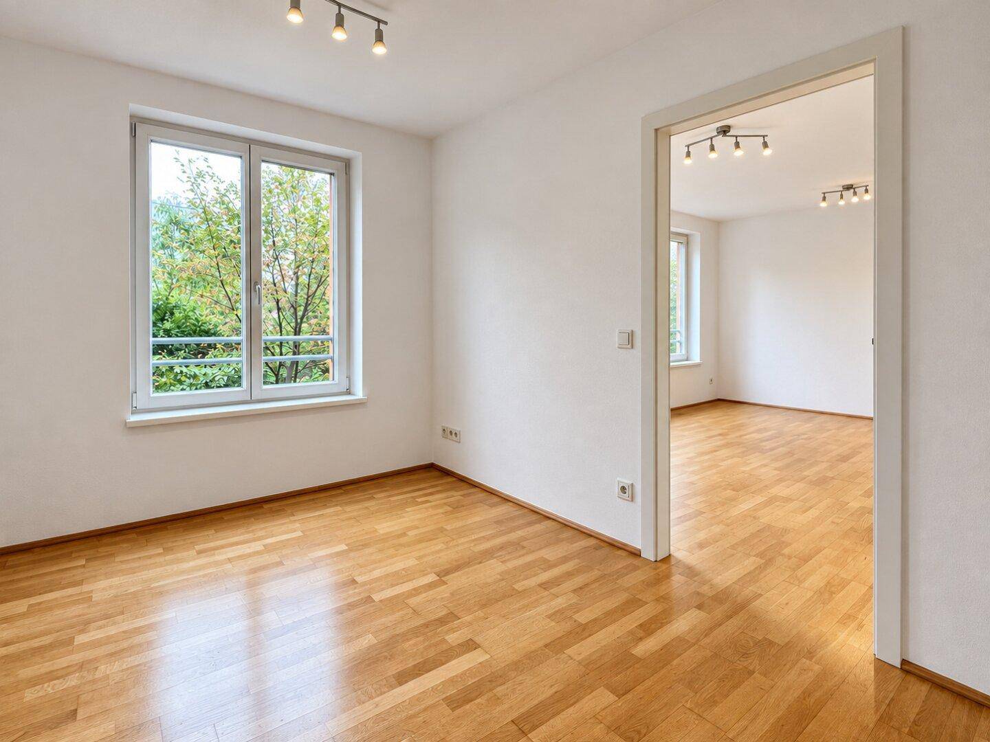 Geräumiger, heller Raum mit Parkettboden und einem Durchgang zu einem weiteren Zimmer.