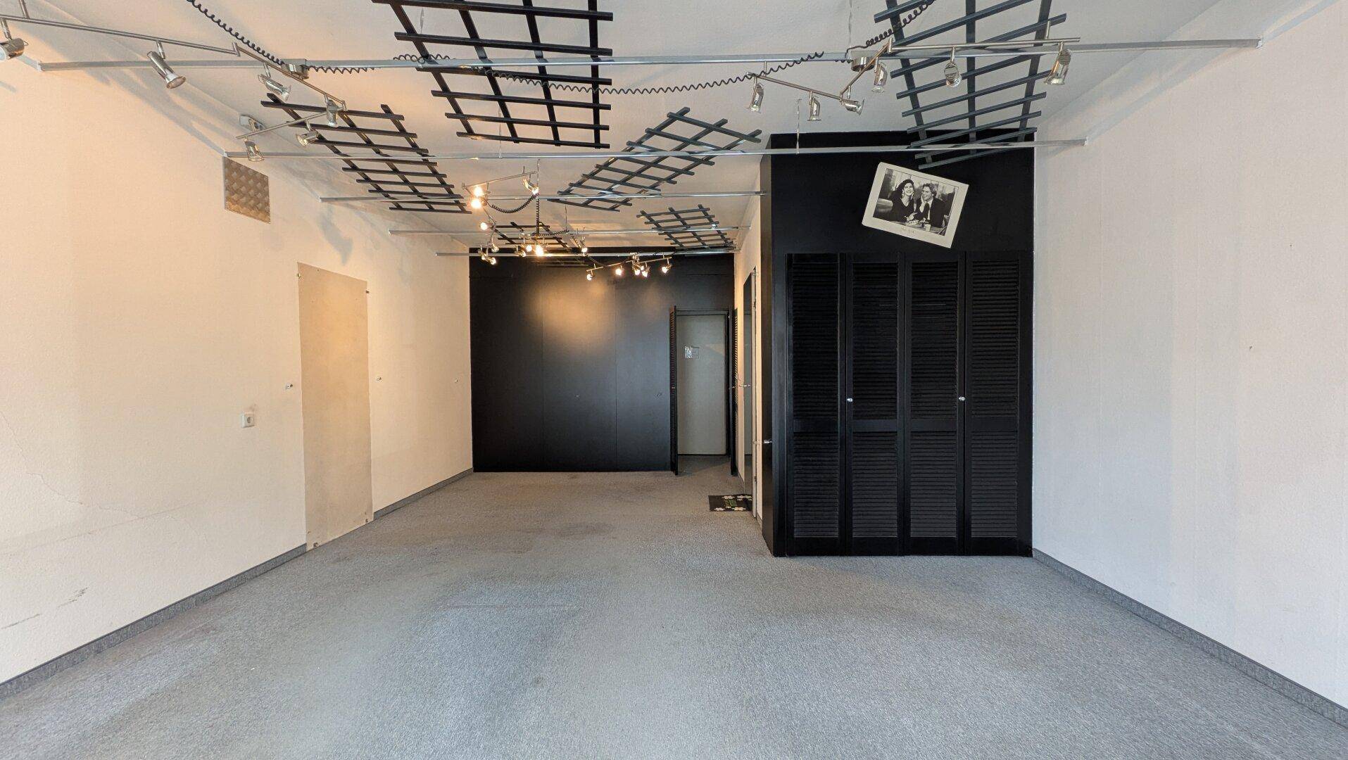 Geräumiger, leerer Ausstellungsraum mit Teppichboden und schwarzen Lamellentüren, ideal für flexible Nutzung.