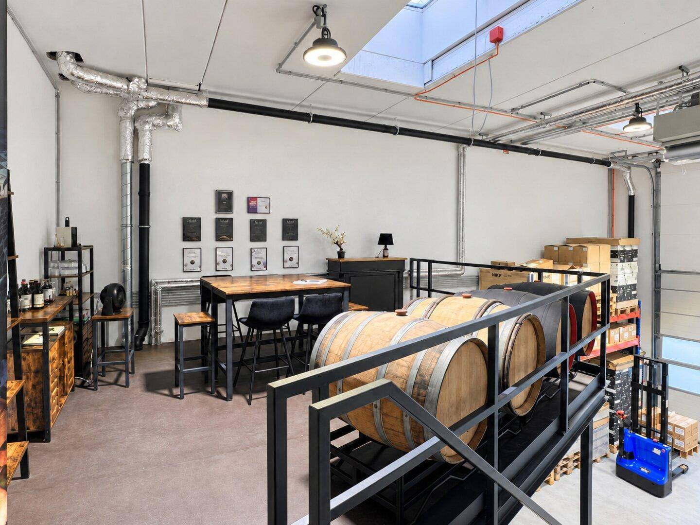 Gemütlicher Loungebereich auf der Galerie mit Holzfässern, Stehtisch und gerahmten Auszeichnungen an der Wand.