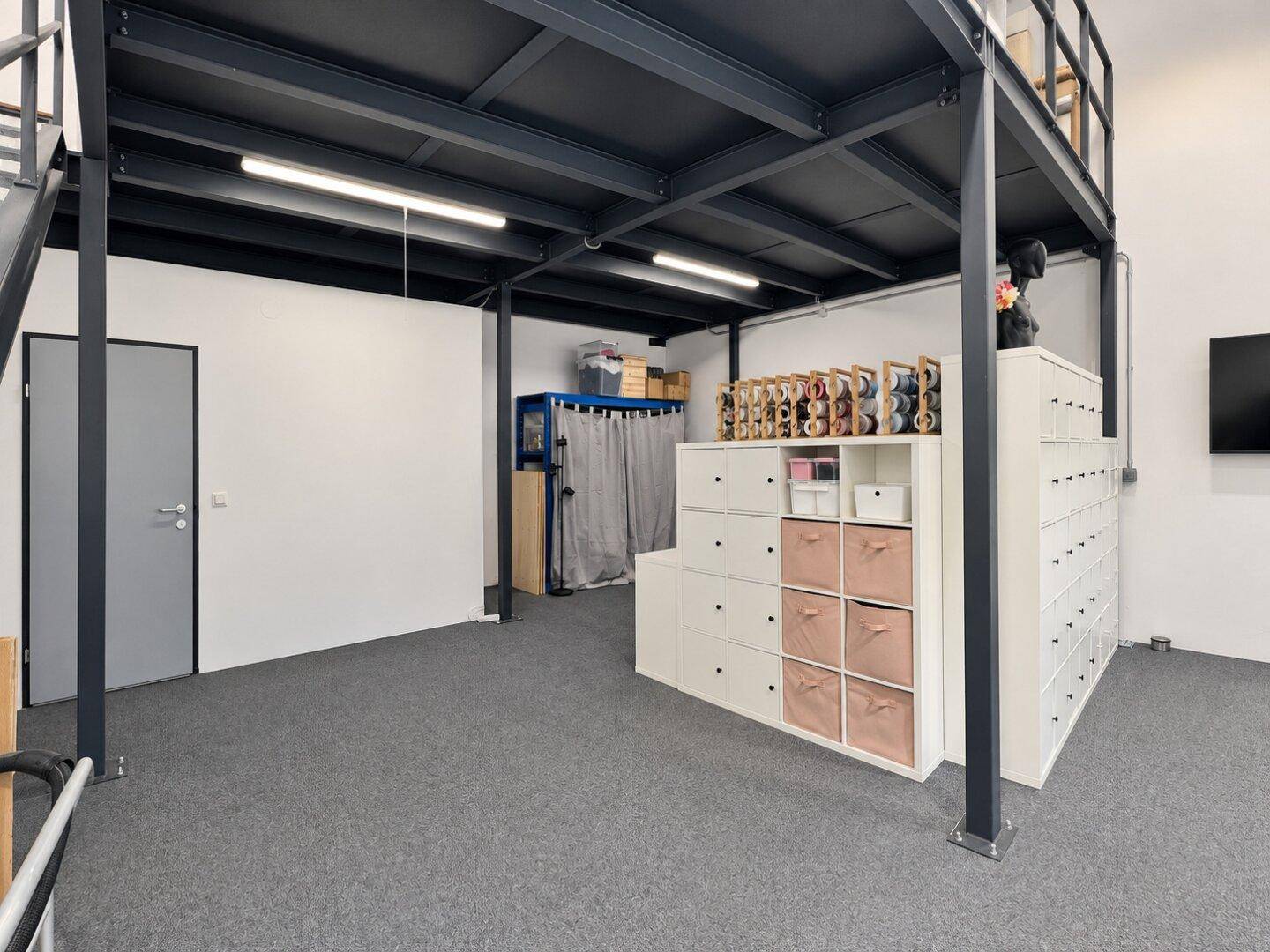 Lagerbereich unter der Galerie mit weißen Regalen, rosa Aufbewahrungsboxen und grauem Teppichboden.