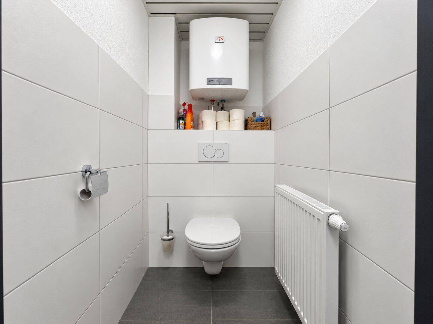 Saubere Toilette mit weißen Fliesen, grauem Boden, wandhängendem WC und einem Heizkörper.