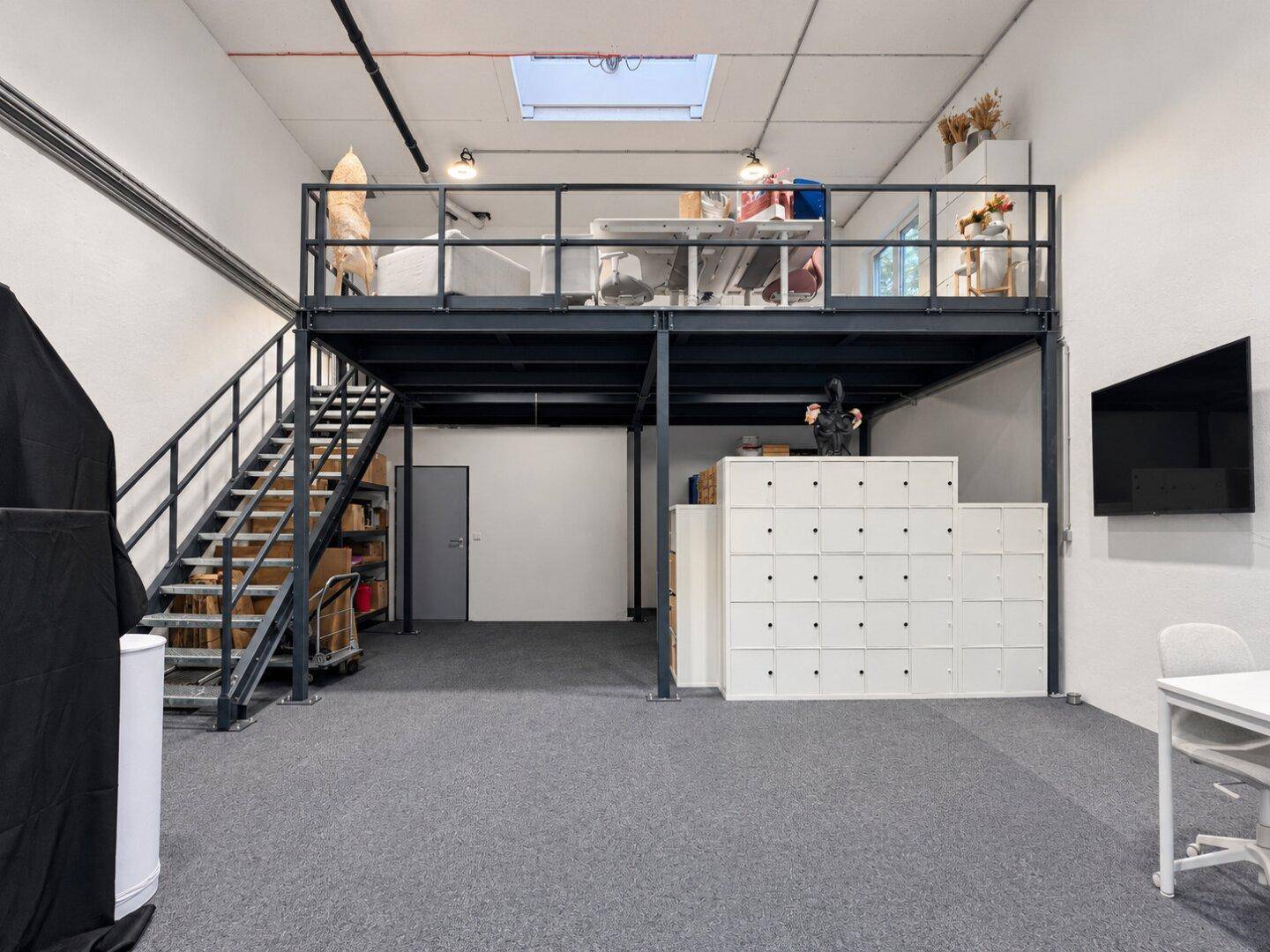 Großer offener Büroraum mit Galerieebene, grauem Teppichboden und weißen Schließfächern.