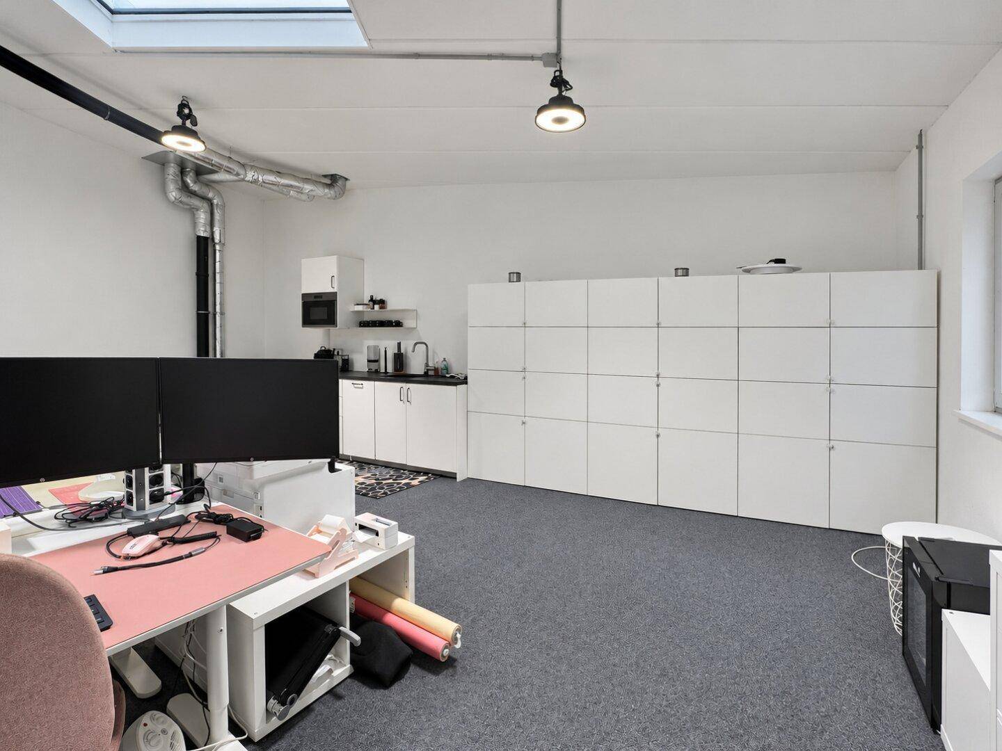 Büroraum mit kleiner Küchenzeile, weißen Schließfächern und Arbeitsplätzen mit Monitoren.