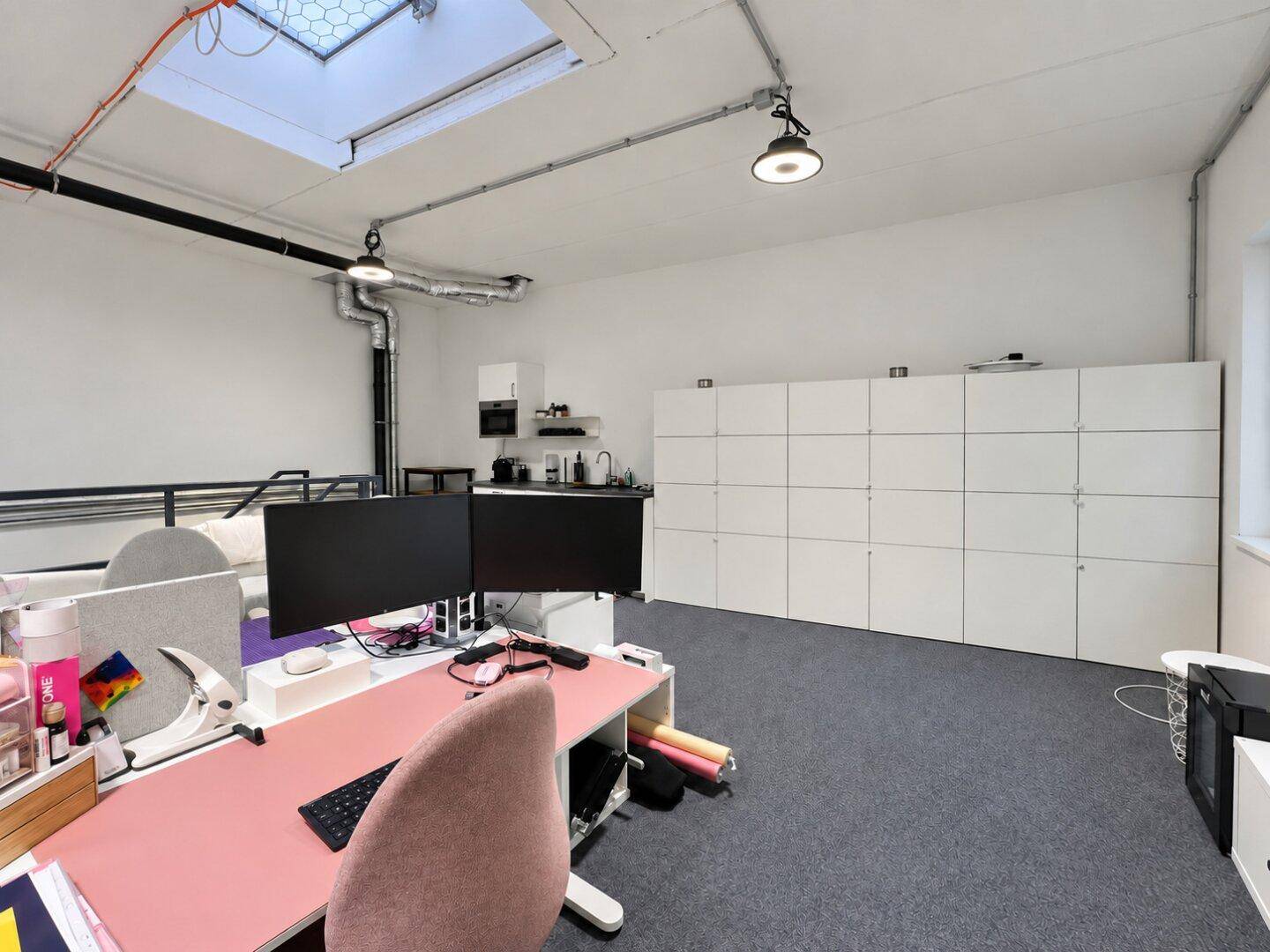 Moderner Büroraum mit Küchenzeile, Schließfächern und einem Arbeitsplatz mit rosa Schreibtisch.
