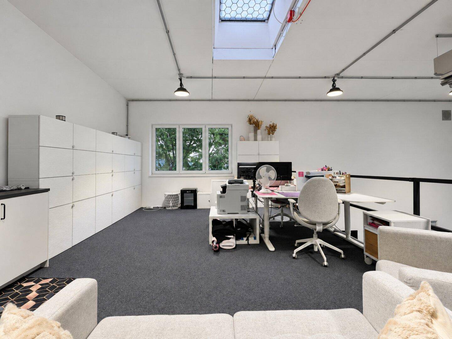 Geräumiger Bürobereich mit weißen Schließfächern, Schreibtischen und einem Sofa, mit Blick ins Grüne.