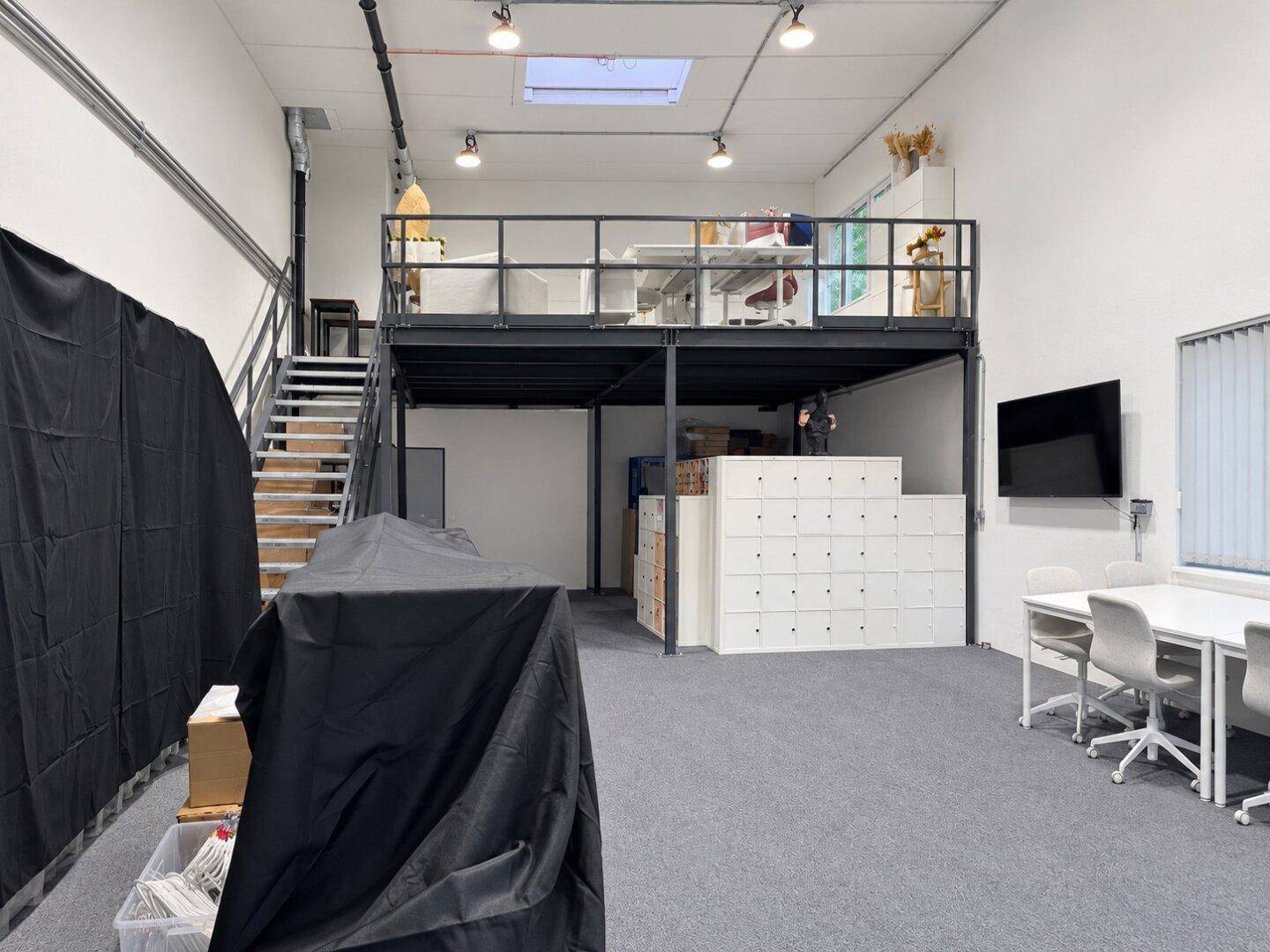 Heller Bürobereich mit Teppichboden, hohen Decken und einer Galerieebene, die zusätzlichen Arbeitsplatz bietet.