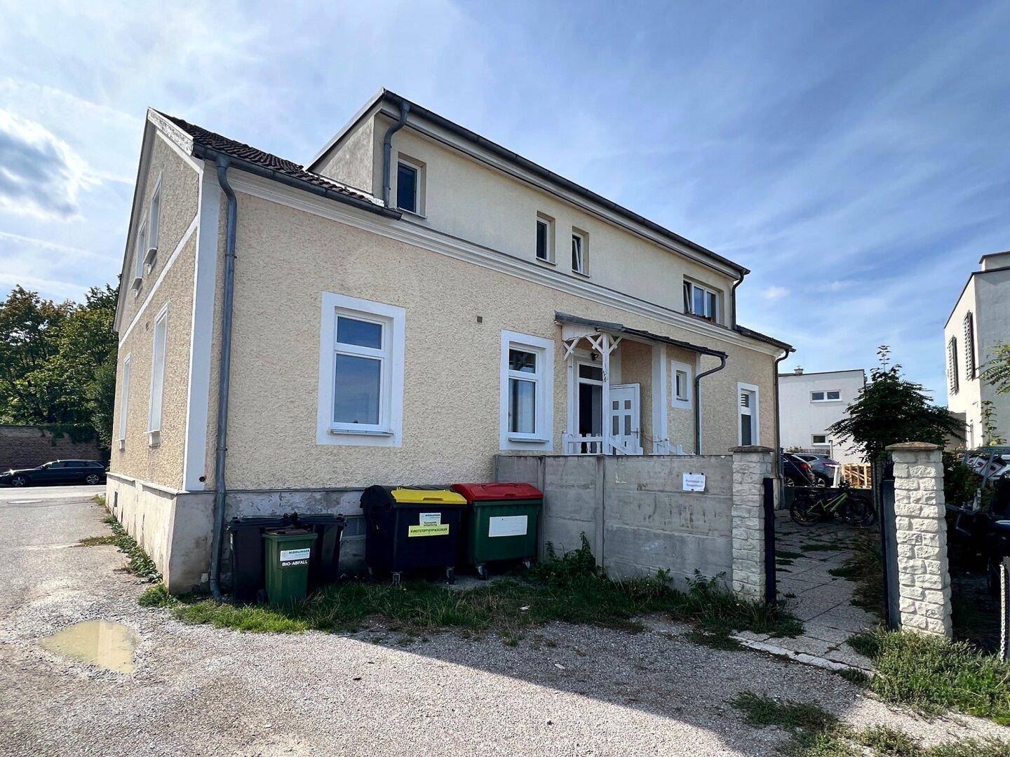 Rückseite des gelben Hauses mit Mülltonnen und einem einfachen Zaun in ländlicher Umgebung.