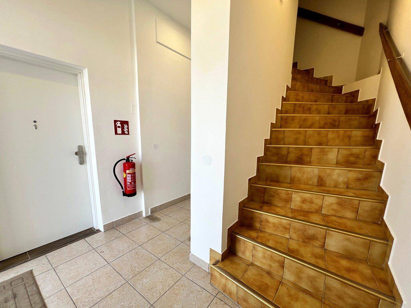 Heller Eingangsbereich mit gefliestem Boden und einer Holztreppe, ausgestattet mit einem Feuerlöscher.