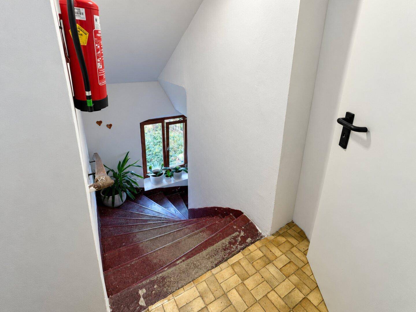 Wendeltreppe mit roten Stufen, weiß gestrichenen Wänden und einem Fenster mit Grünblick.