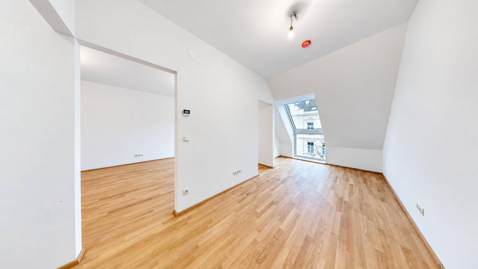 Leeres Zimmer mit Holzboden, weiß gestrichenen Wänden und einem Dachfenster.