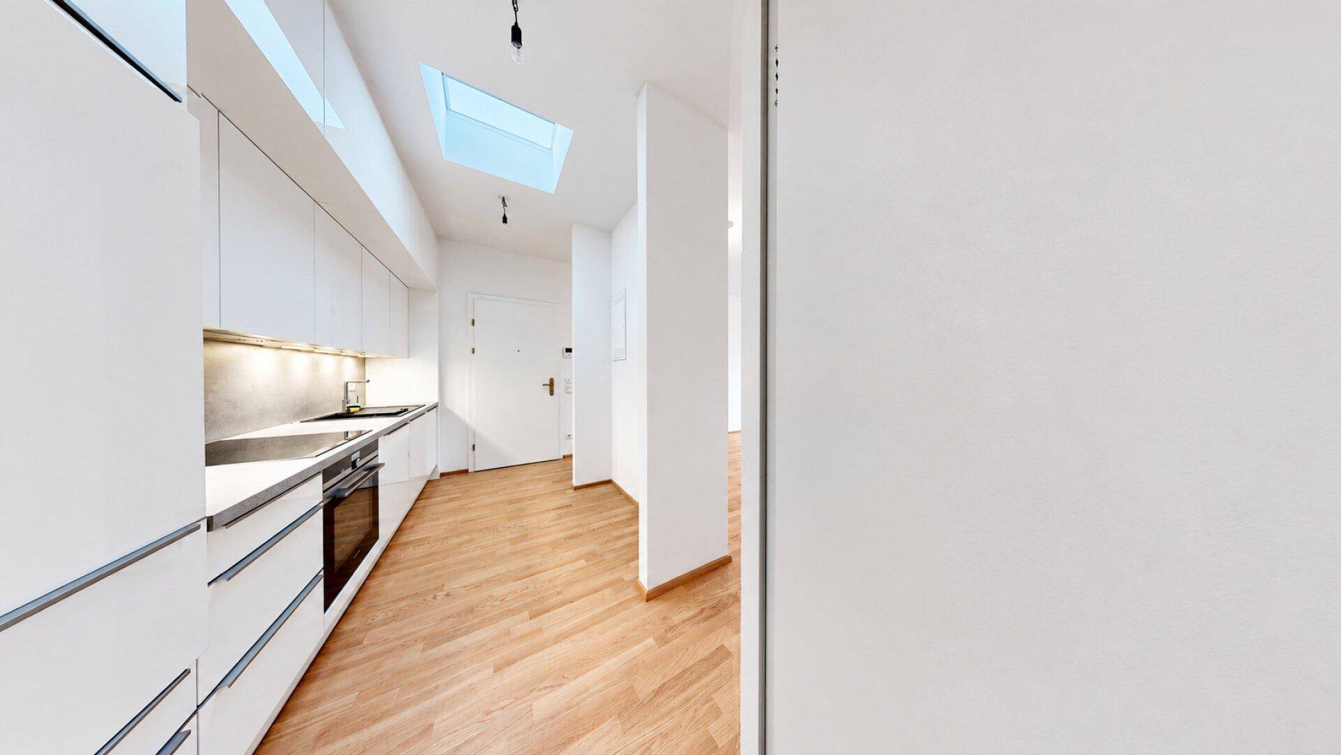 Heller Küchenbereich mit modernen weißen Schränken und Zugang zum Flur, beleuchtet durch ein Dachfenster.