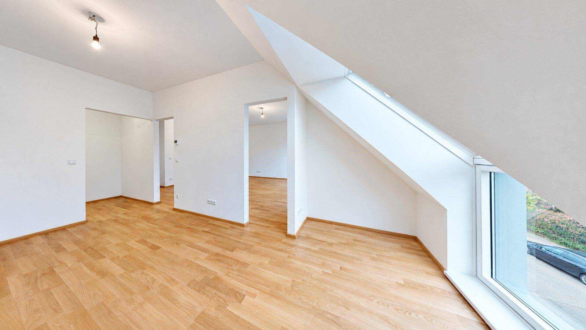 Helles Zimmer mit Holzboden und einer großen, schrägen Fensterfront, die viel Licht hereinlässt.