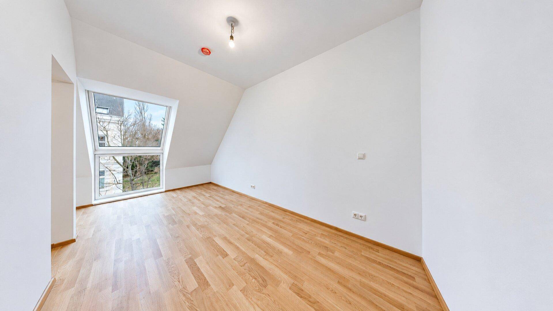 Geräumiges, helles Zimmer mit Holzboden und großer Dachgaube, ideal als Schlaf- oder Wohnzimmer.