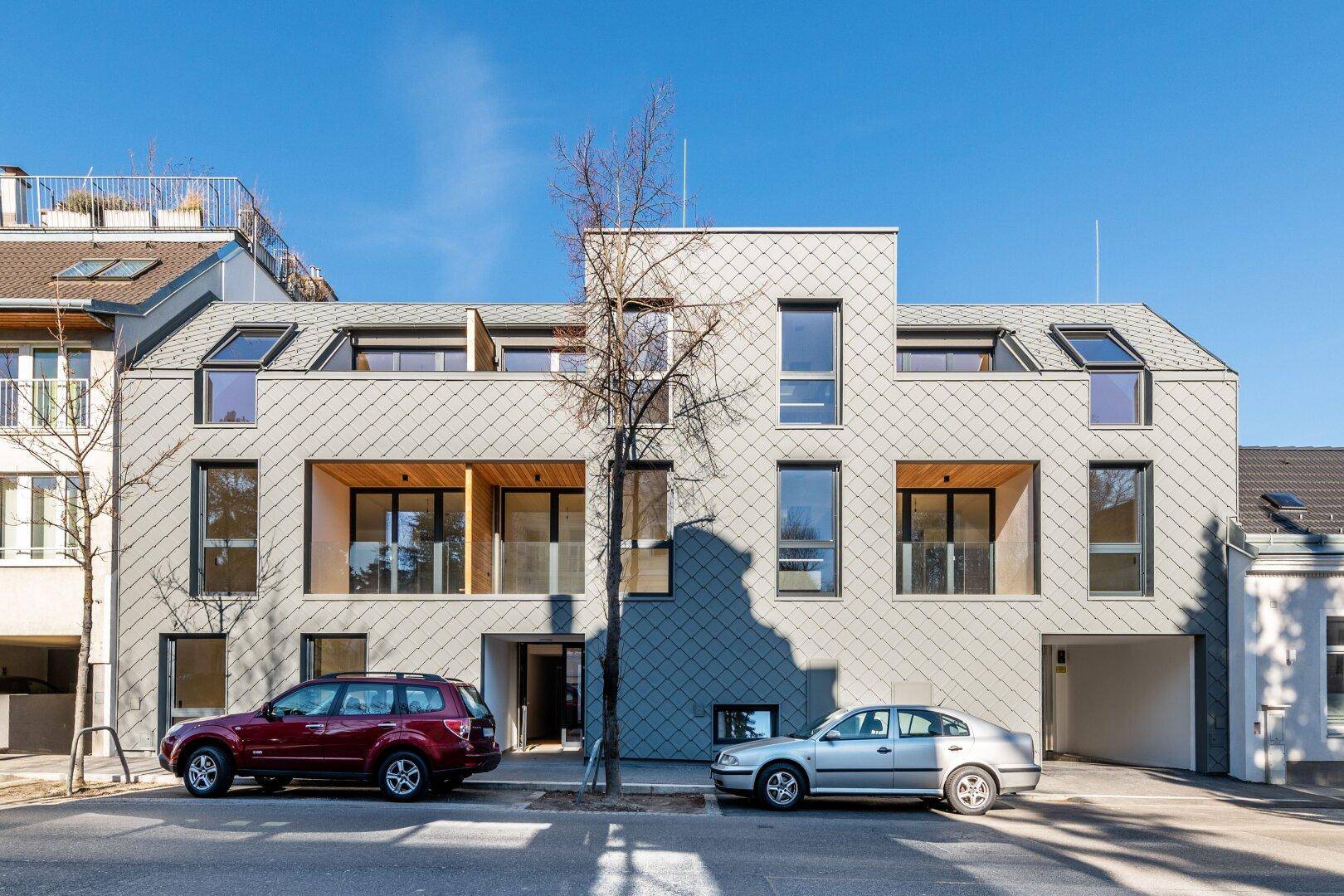 Moderne Wohngebäude-Fassade mit Schindeln, großen Fenstern und Parkplätzen davor.