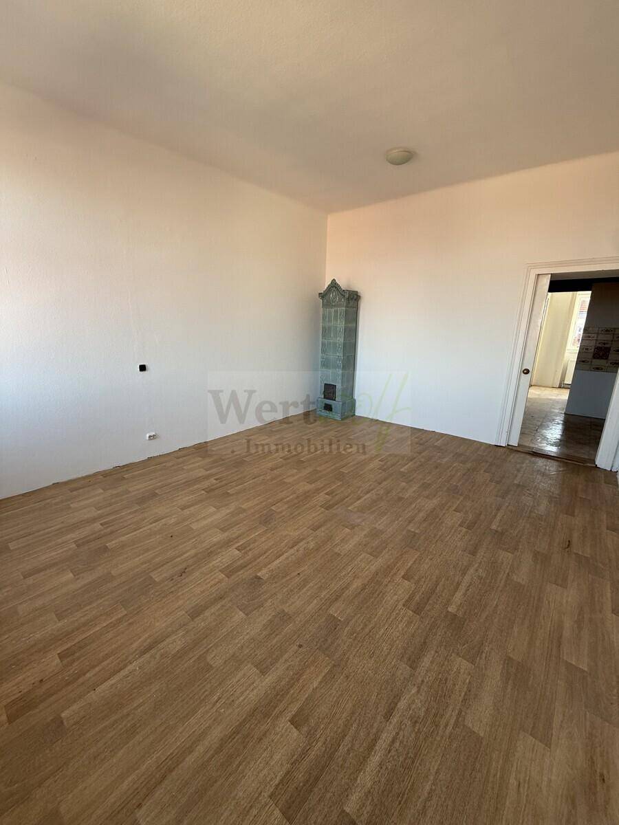 Geräumiges Zimmer mit hellem Laminatboden und einem traditionellen Kachelofen, bereit zur Renovierung.