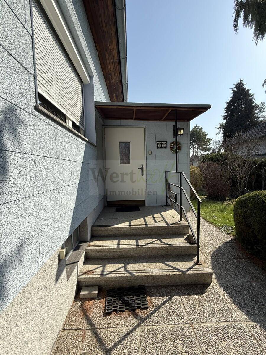 Überdachter Hauseingang mit Treppe, Geländer und einem Fenster mit Rollladen.