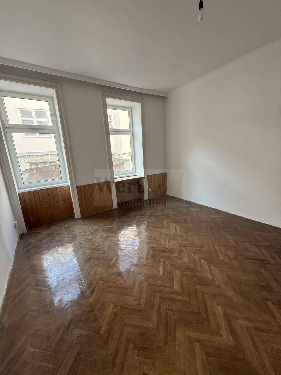 Geräumiges Zimmer mit Fischgrätparkett, hohen Fenstern und einer Holzvertäfelung im unteren Bereich.