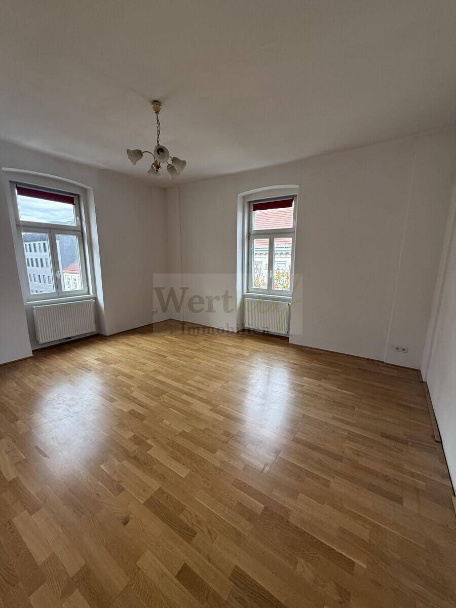 Helles Zimmer mit klassischem Parkettboden und zwei Fenstern, ausgestattet mit einem Deckenleuchter.
