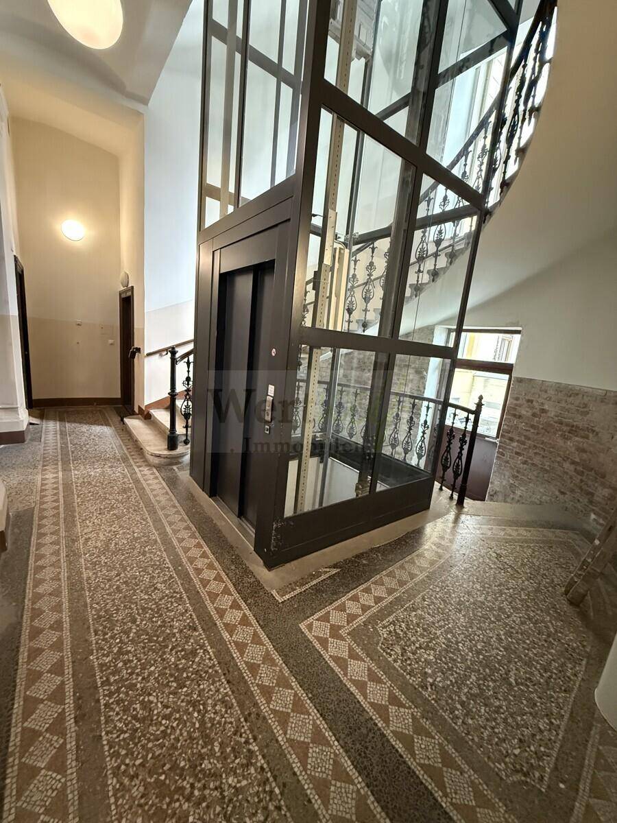 Treppenhaus mit modernem Glasaufzug und historischem Geländer.