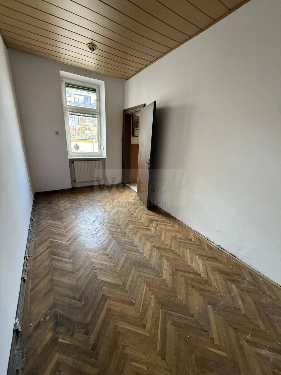 Kleinerer Raum mit Fischgrätparkett, Holzdecke und einem Fenster zur Straße, das Licht hereinlässt.