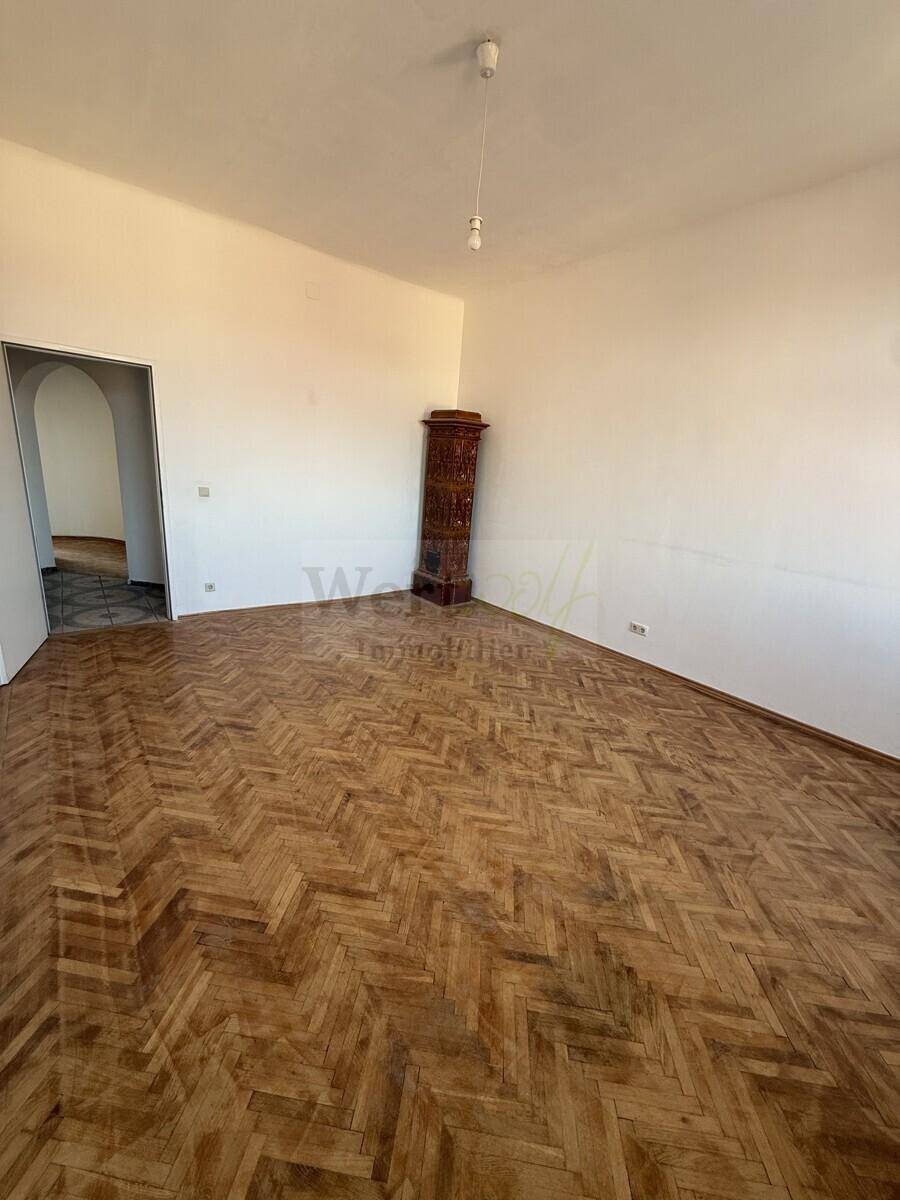 Geräumiges Zimmer mit Fischgrätparkett und einem traditionellen Kachelofen, bereit zur Renovierung.
