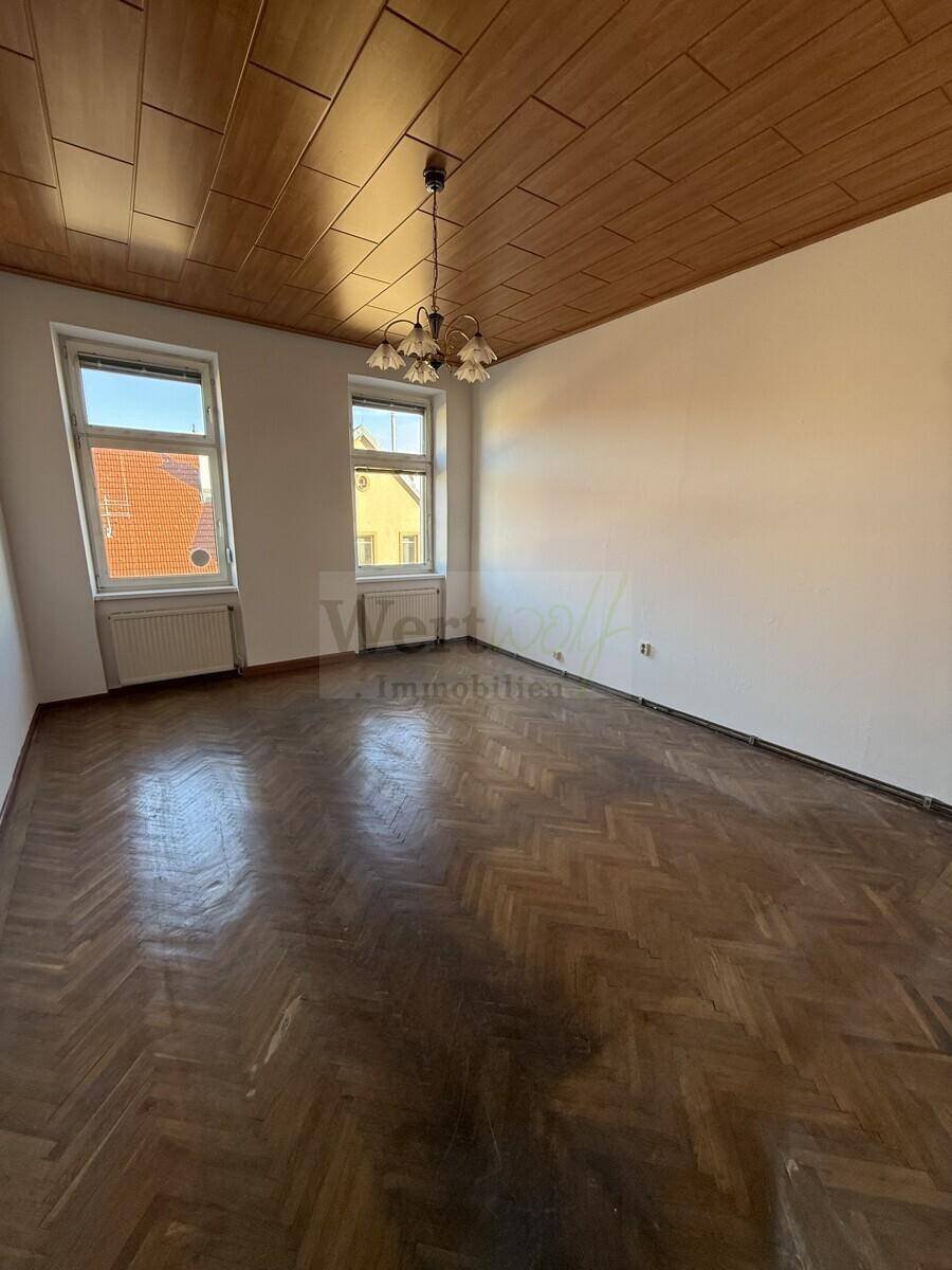 Geräumiger Raum mit Fischgrätparkett, Holzdecke und zwei großen Fenstern, bereit zur Renovierung.