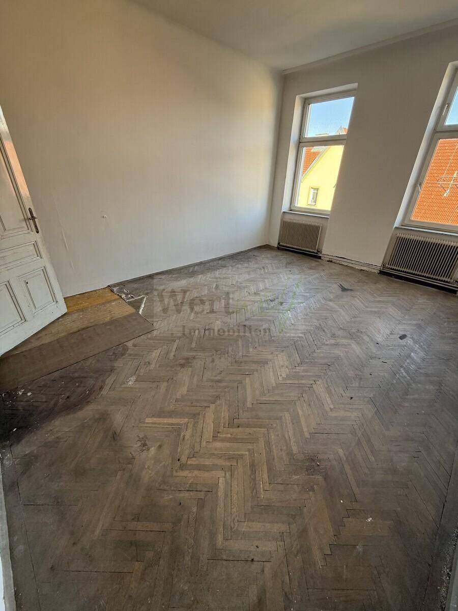 Großer Raum mit abgenutztem Fischgrätparkett und zwei Fenstern, der eine umfassende Renovierung benötigt.