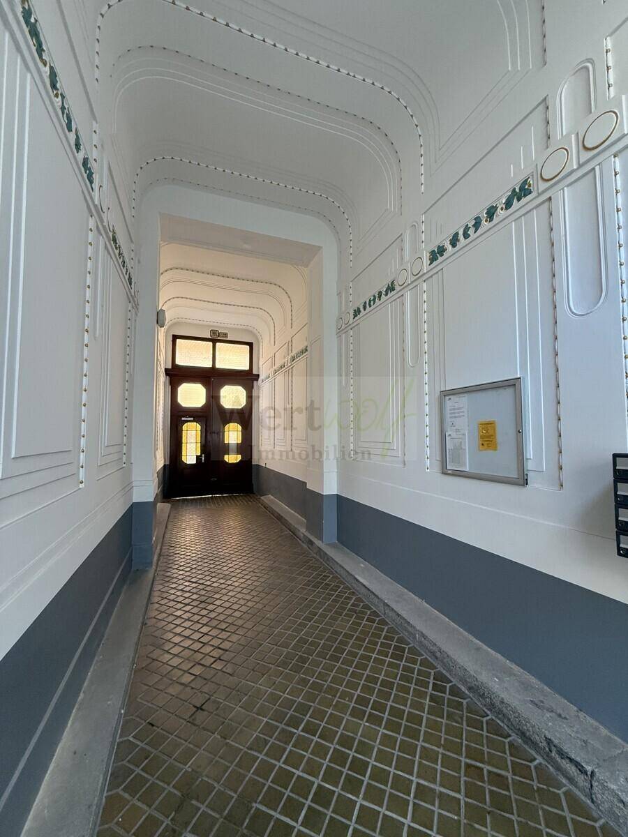 Langer, heller Eingangsbereich mit dekorativen Wandpaneelen und gemustertem Fliesenboden, der zum Hauptgebäude führt.