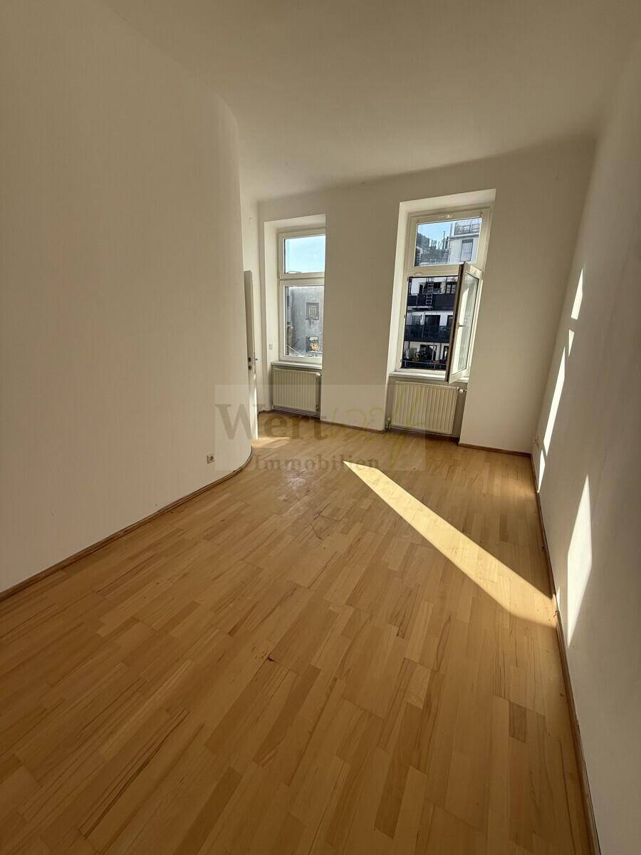 Sonniges Zimmer mit hellem Holzparkett und zwei Fenstern, die viel Tageslicht hereinlassen.