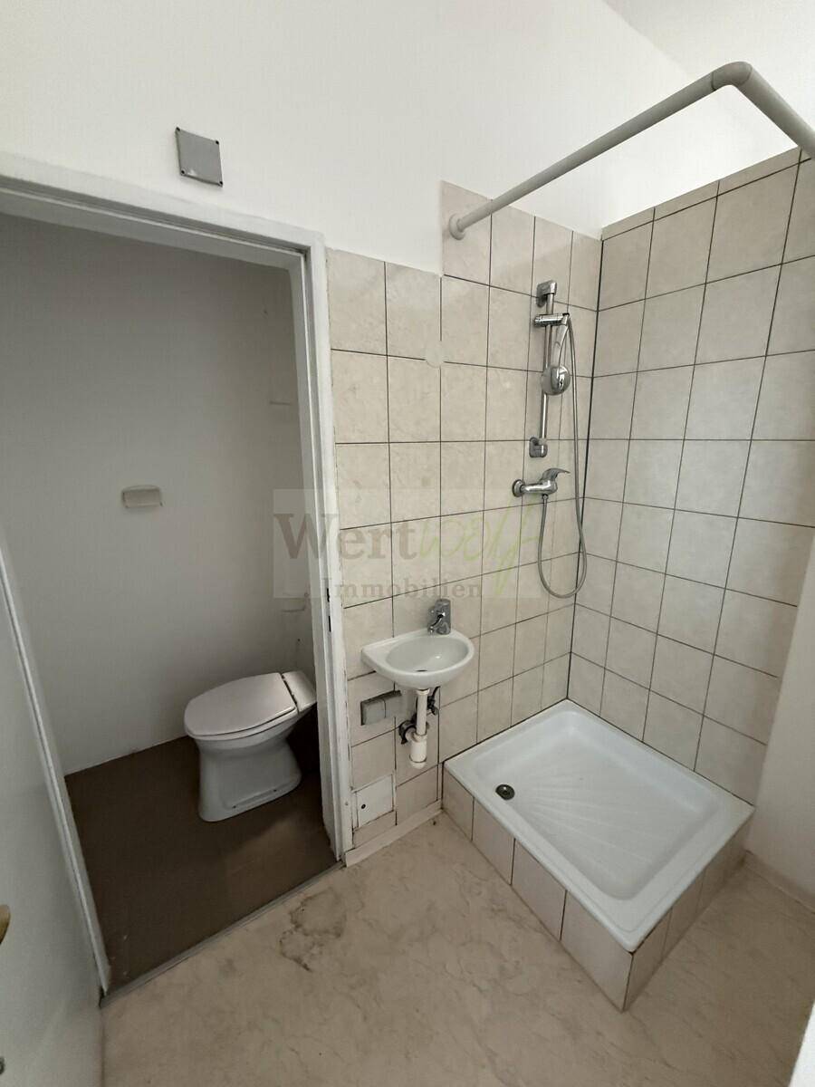 Einfaches Badezimmer mit Duschwanne, WC und Waschbecken, renovierungsbedürftig.