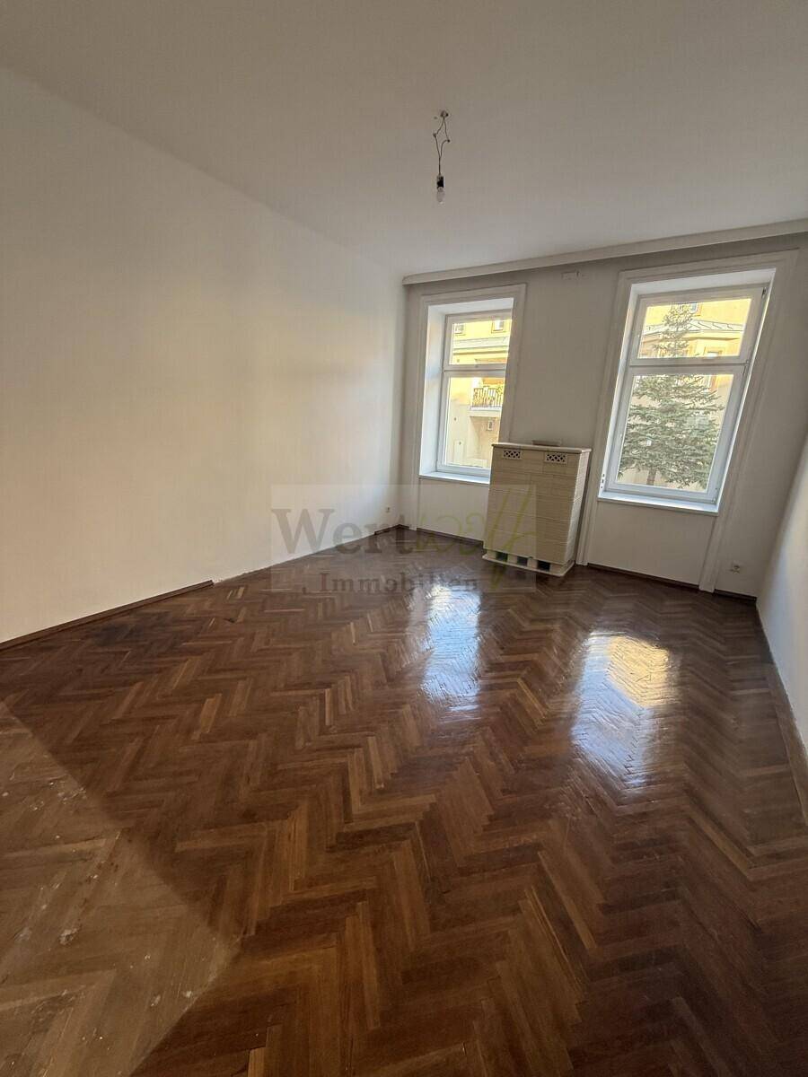 Lichtdurchflutetes Zimmer mit elegantem Fischgrätparkett und Blick auf die Umgebung durch hohe Fenster.