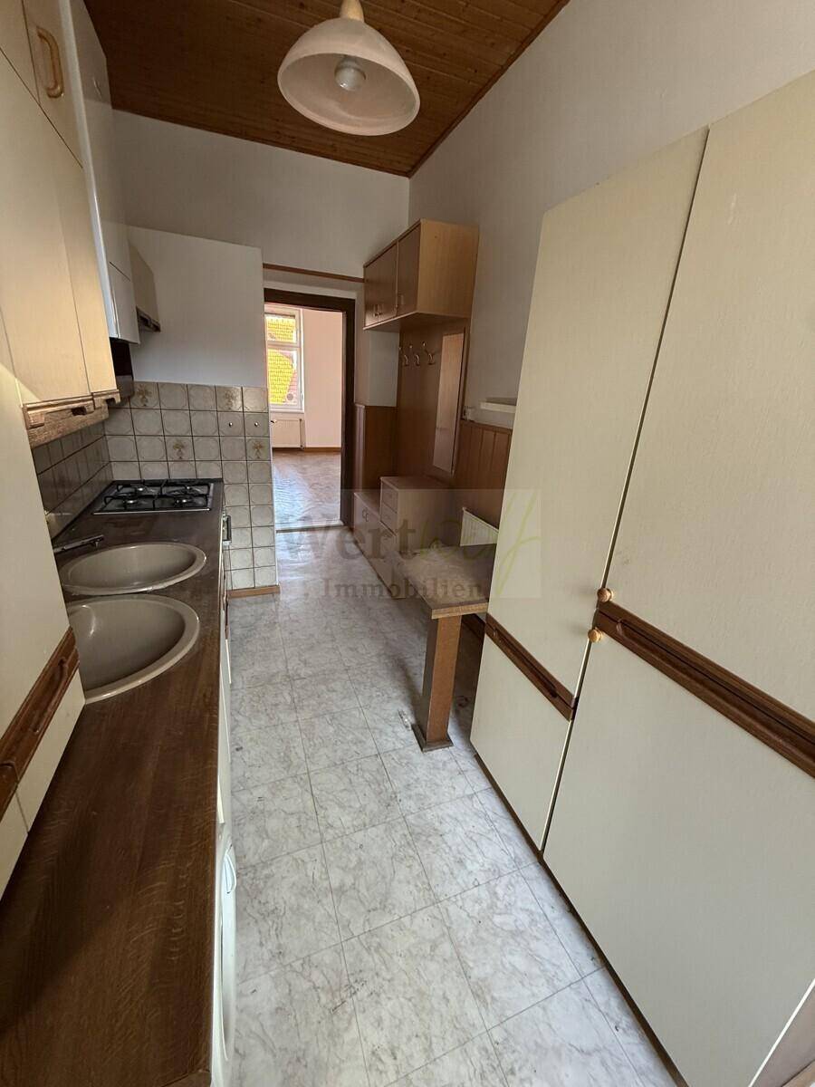 Schmale Küche mit älteren Einbaumöbeln, Spüle und Blick in den Flur, der renoviert werden muss.