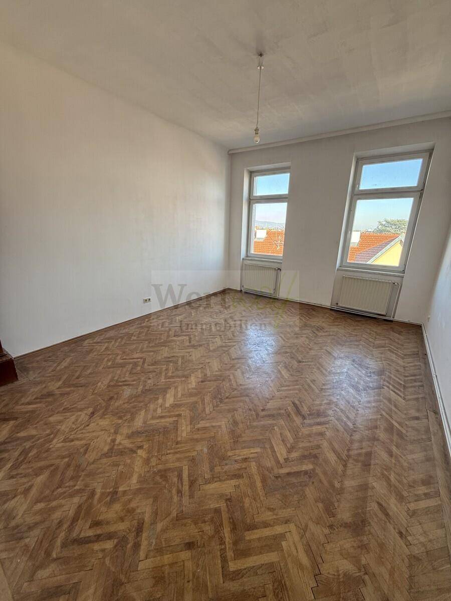 Helles Zimmer mit Fischgrätparkett, zwei Fenstern und Blick auf die Dächer der Stadt.