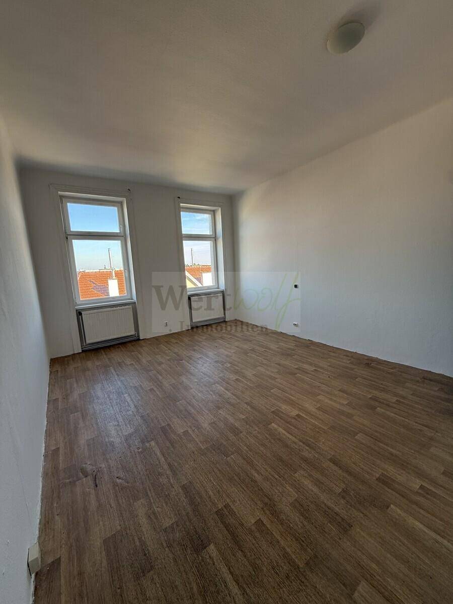 Helles Zimmer mit zwei Fenstern, Laminatboden und Heizkörpern unter den Fenstern.