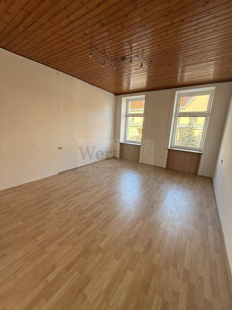 Großes Zimmer mit zwei Fenstern, Parkettboden und einer Holzdecke, die eine warme und einladende Atmosphäre schafft.