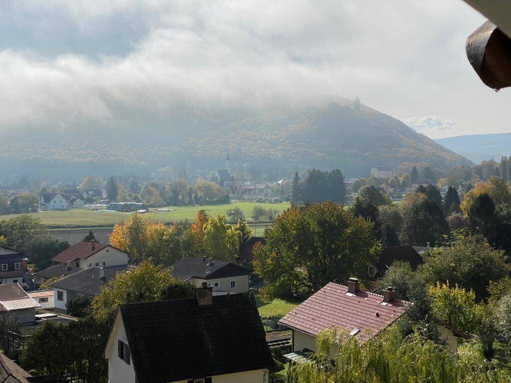 Weitläufiger Ausblick über eine Siedlung, grüne Felder und bewaldete Berge unter bewölktem Himmel.