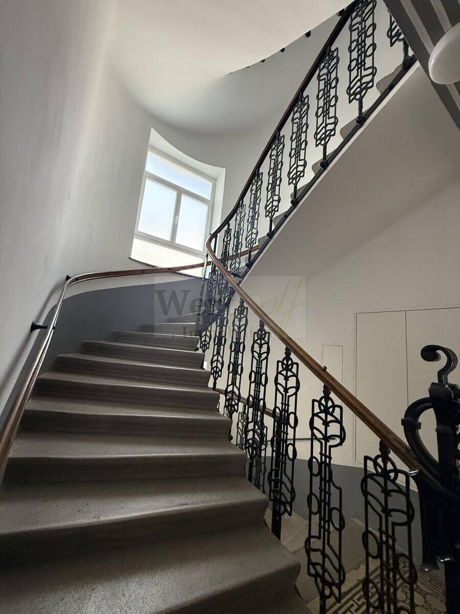 Historisches Treppenhaus mit schmiedeeisernem Geländer, Steinstufen und natürlichem Lichteinfall durch ein Fenster.