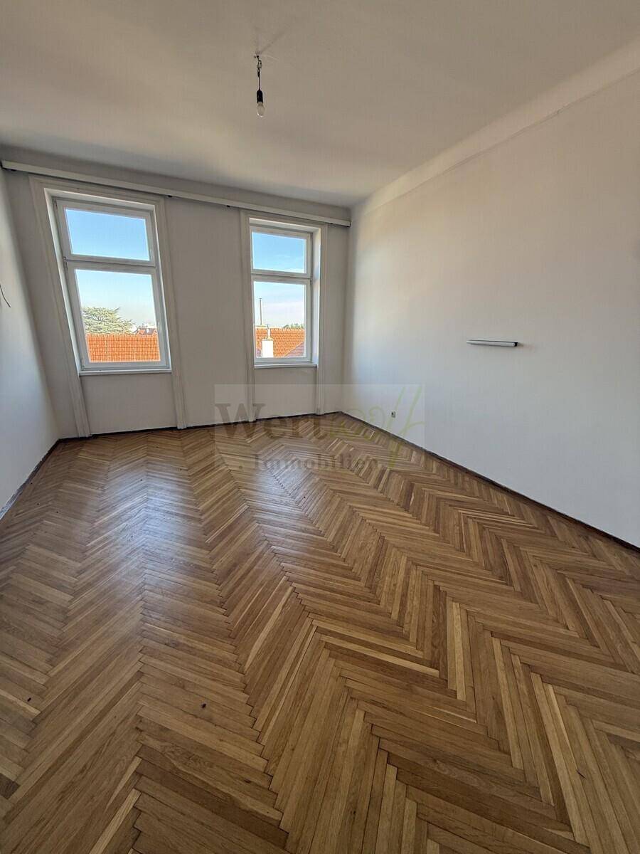 Geräumiges Zimmer mit Fischgrätparkettboden und zwei Fenstern, die viel Tageslicht hereinlassen.