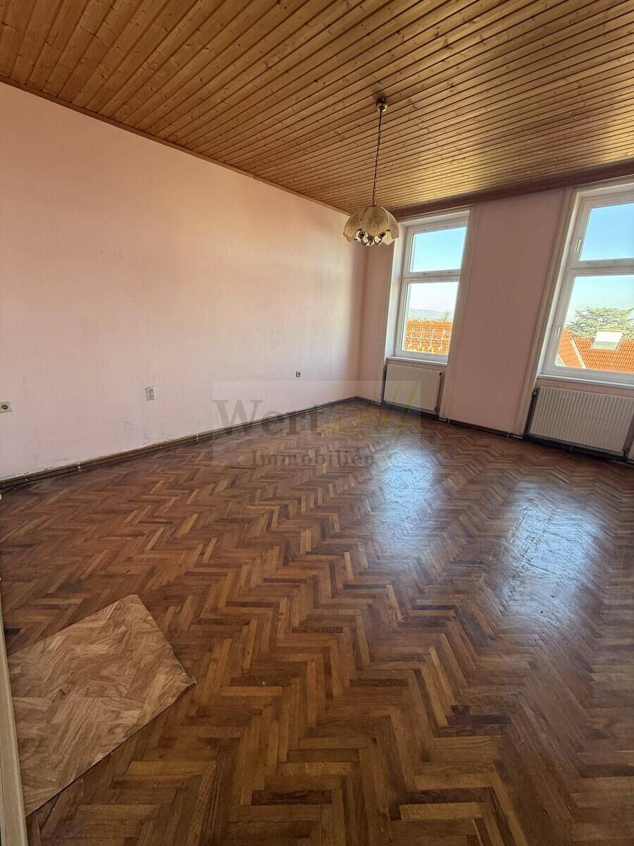 Geräumiges Zimmer mit Fischgrätparkett, Holzdecke und zwei Fenstern, die den Raum erhellen.