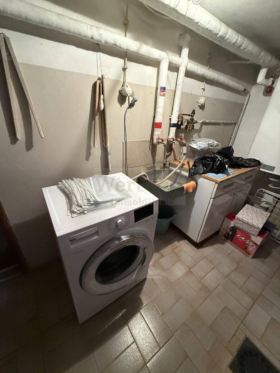 Waschküche im Keller mit Waschmaschine, Spülbecken und sichtbaren Rohren.
