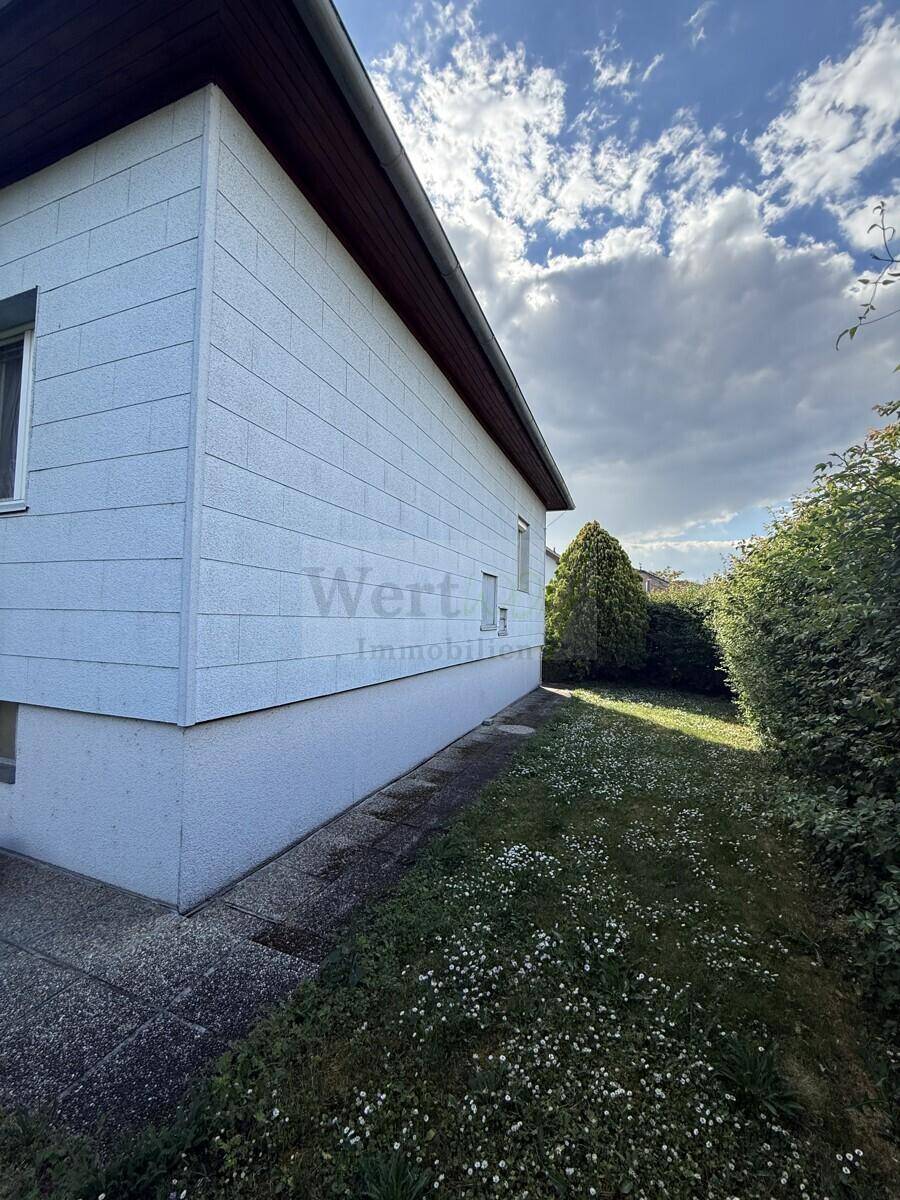 Seitenansicht des Hauses mit heller Fassade und einem schmalen Grünstreifen entlang des Gebäudes.