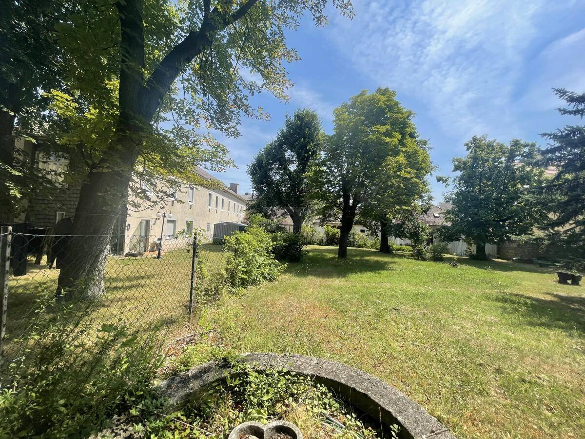 Gartenfläche mit Zaun und altem Baumbestand, angrenzend an ein längliches Wohngebäude.