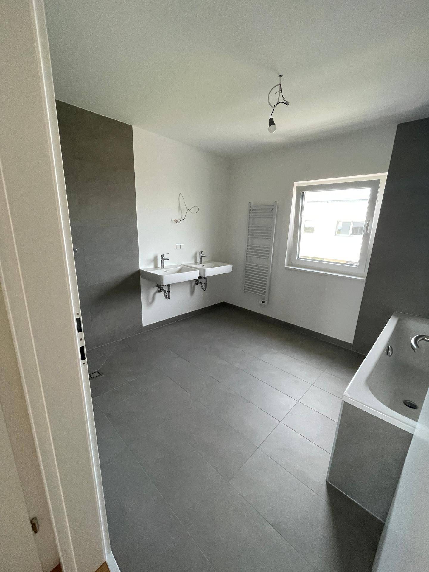 Modernes Badezimmer mit grauen Fliesen, Doppelwaschbecken, Badewanne und praktischem Handtuchheizkörper.