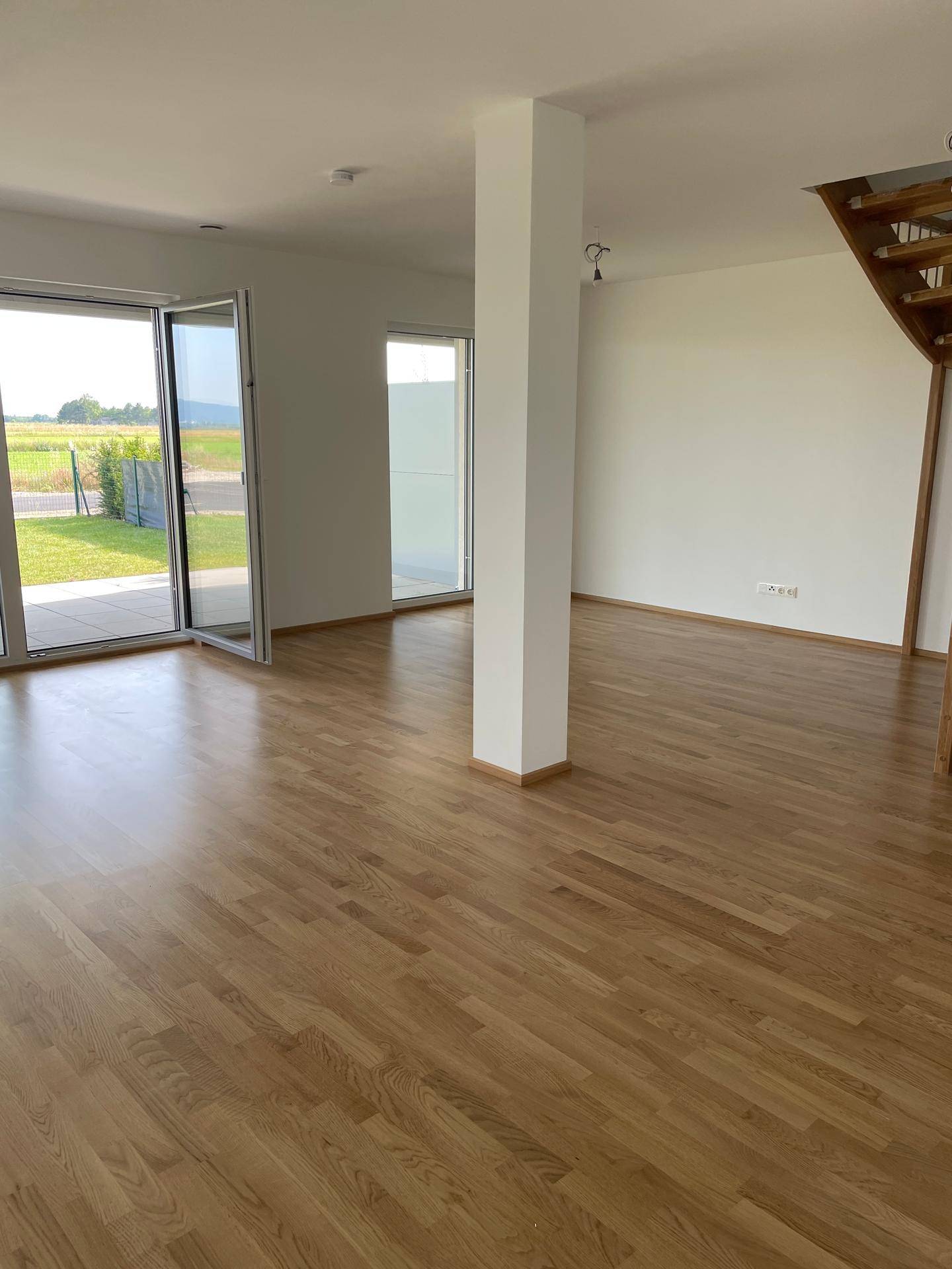 Offene Wohnfläche mit Holzboden, einem zentralen Pfeiler und großen Fenstern zum Außenbereich.