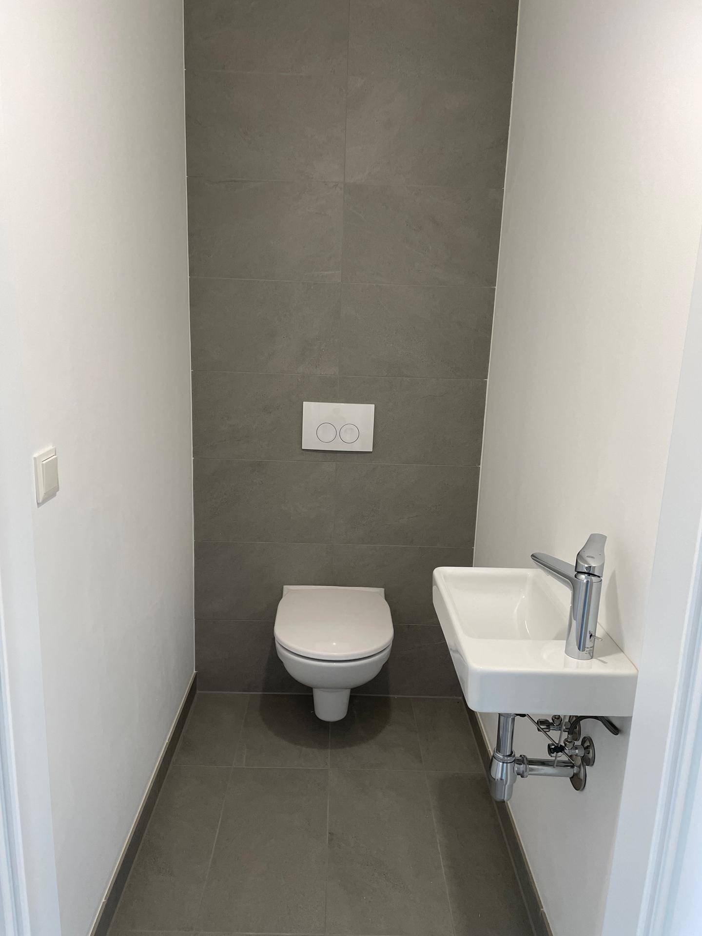 Modernes Gäste-WC mit grauen Fliesen, wandhängendem WC und einem kleinen, praktischen Waschbecken.