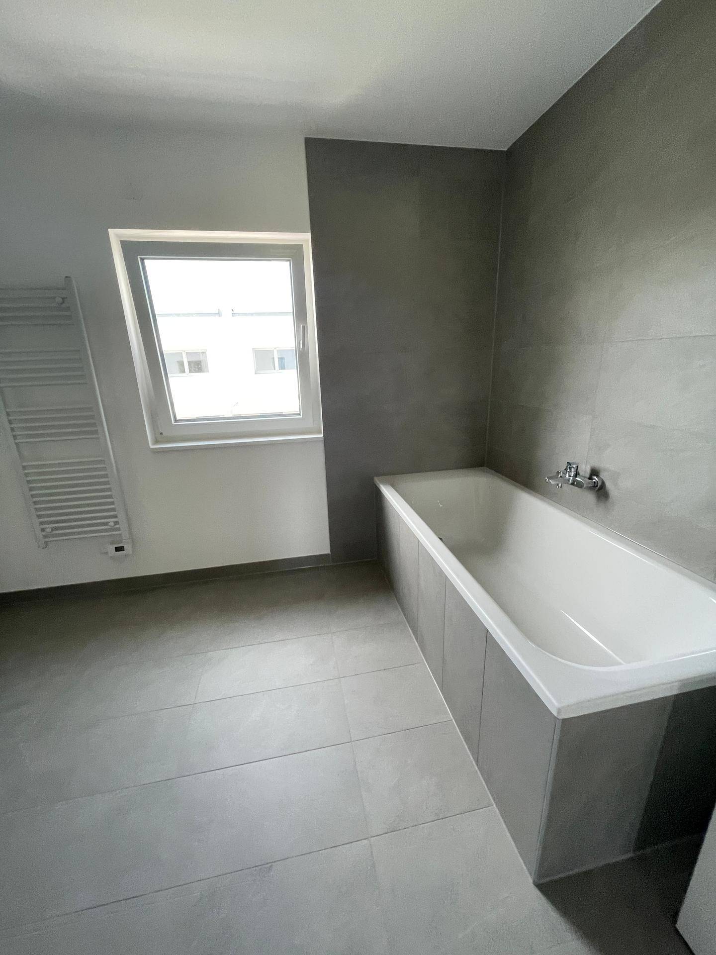 Helles Badezimmer mit Badewanne, grauen Fliesen und einem Fenster für natürliche Belüftung und Licht.