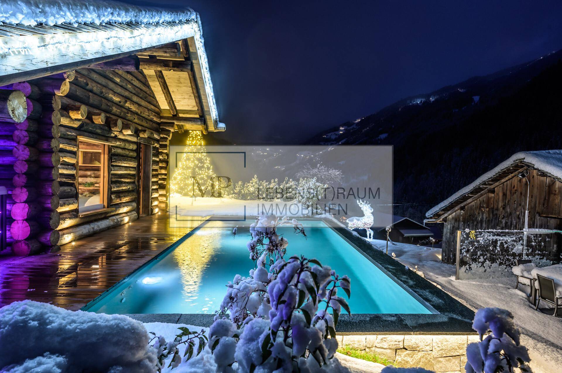 Beleuchteter Außenpool im Schnee vor einer rustikalen Holzhütte mit winterlicher Bergkulisse bei Nacht.