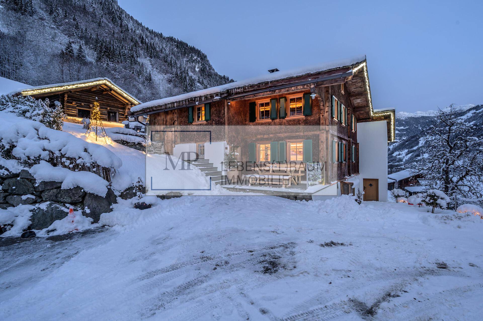Traditionelles Holzchalet in einer verschneiten Berglandschaft bei Dämmerung mit beleuchteten Fenstern.