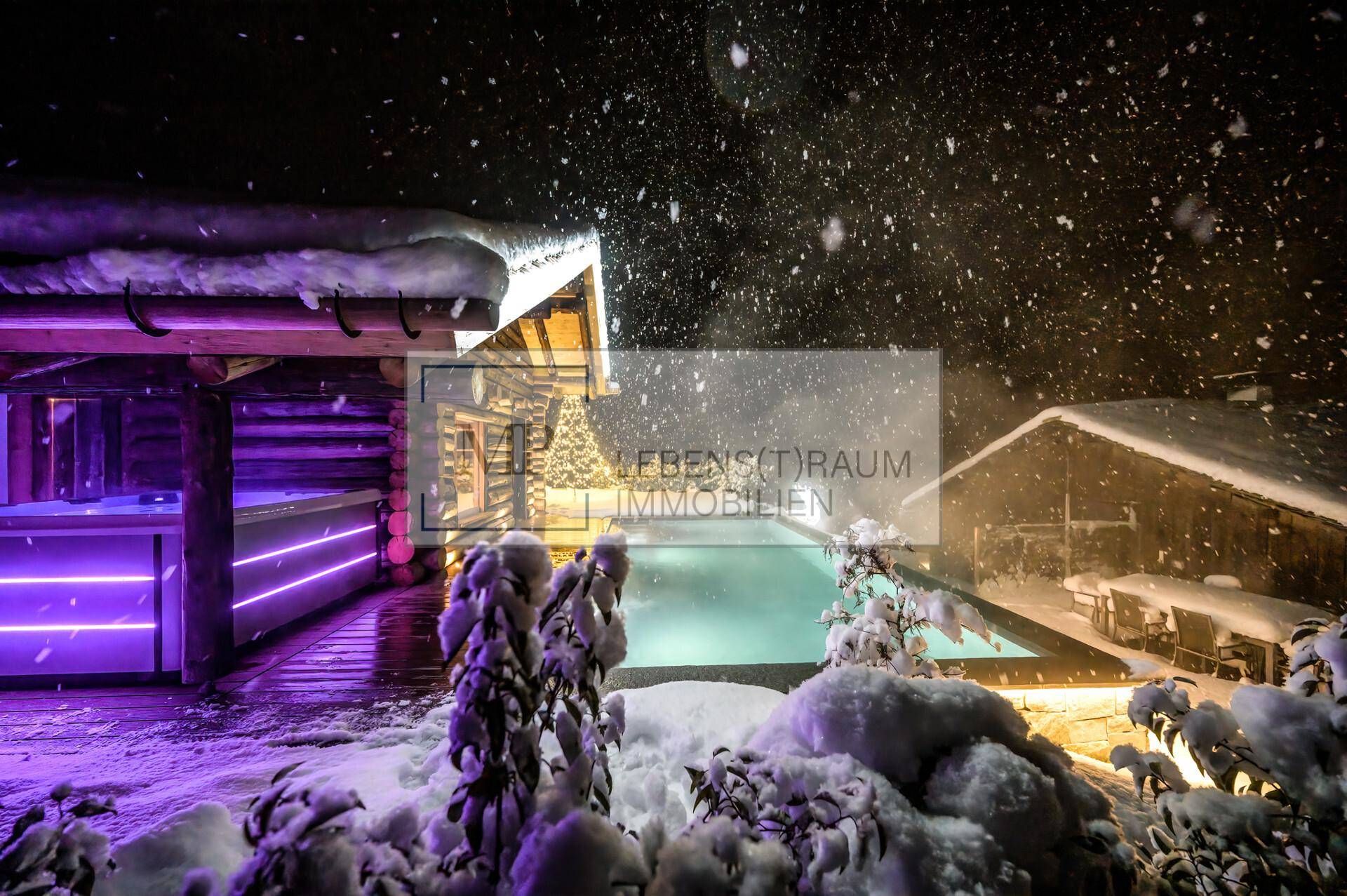 Beleuchteter Außenpool im Schnee mit lila Lichtern vor einer rustikalen Holzhütte und winterlicher Bergkulisse.