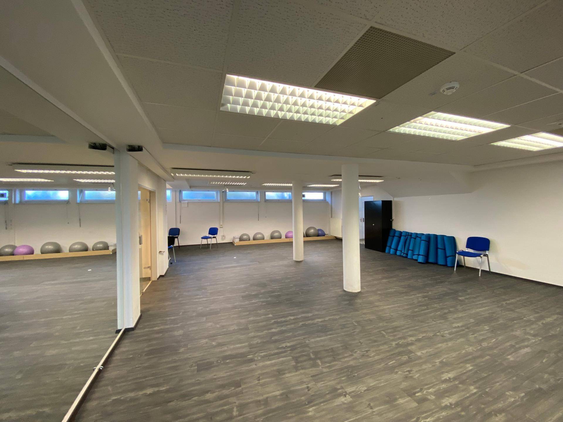 Großer Mehrzweckraum mit dunklem Vinylboden, vielen Fenstern und Säulen, ideal für Sportaktivitäten.